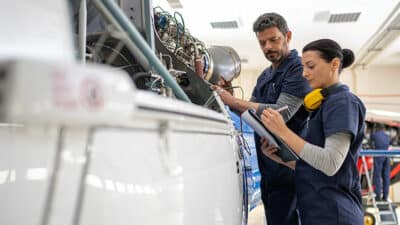 Male and female mechanics with clipboard and voltage tester working on helicopter engine in hangar.