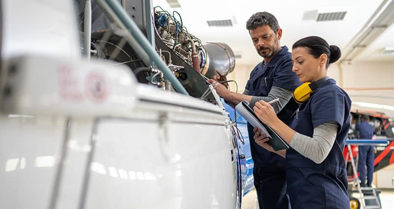 Male and female mechanics with clipboard and voltage tester working on helicopter engine in hangar.